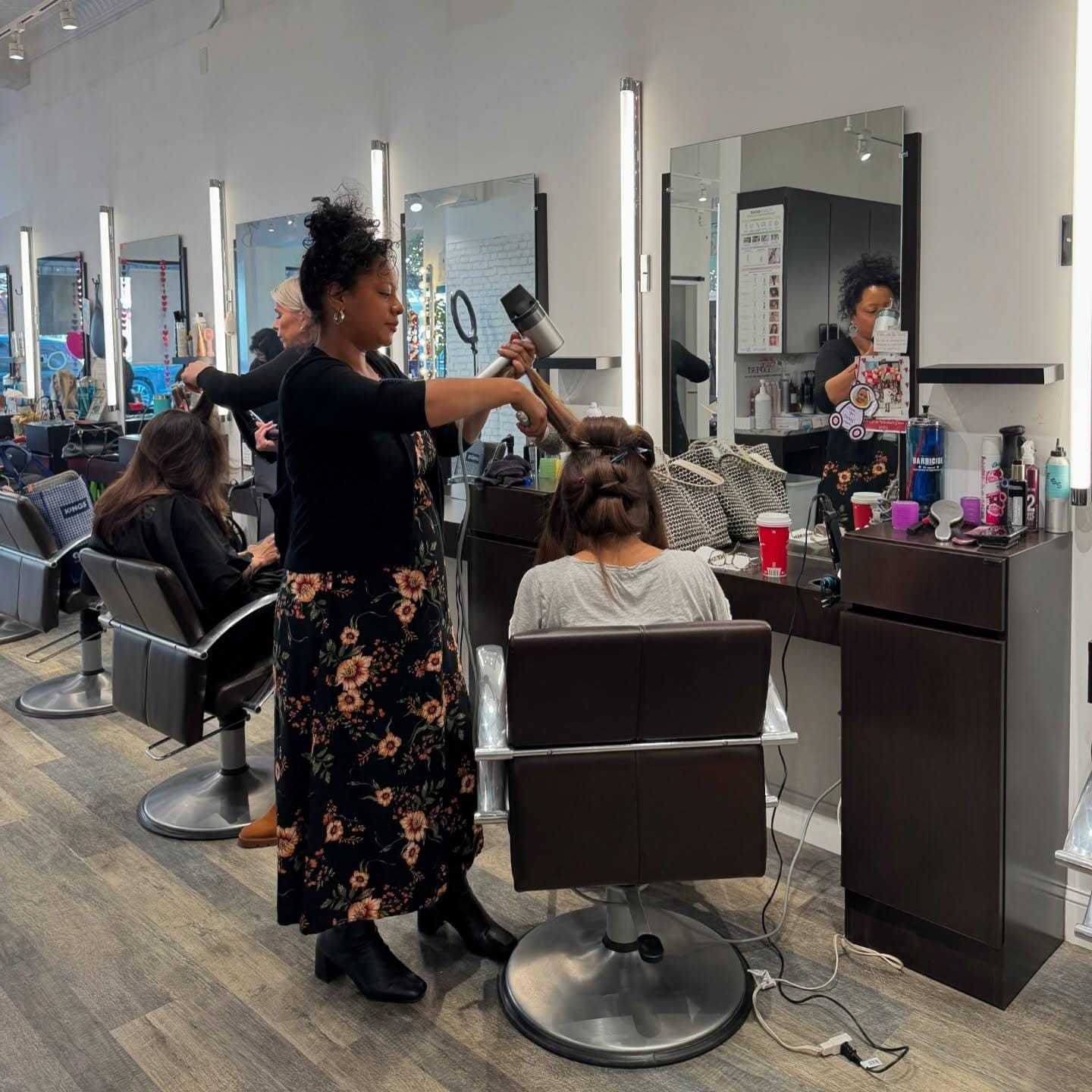 Hair salon interior with a stylist blow-drying a client's hair.